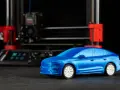 Niebieski model samochodu, gotowy do drukowania, stoi przed drukarką 3D.