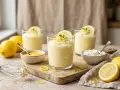 Trois verres de mousse au citron, décorés de rondelles de citron et de menthe. Des citrons frais et des ingrédients pour la préparation sont disposés autour.