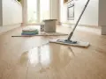 Ein Mopp wischt über einen glänzenden Holzboden. Reinigungsmittel und Tücher liegen bereit, um den **Vinylboden zu reinigen**.