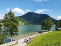 Ein sonniger Tag am bayrischzell see. Menschen spazieren am Ufer entlang, während im Hintergrund ein bewaldeter Berg aufragt.