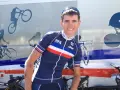 David Gaudu, a French cyclist, smiles in his team's blue, white, and red jersey, with bike graphics on the vehicle behind him.