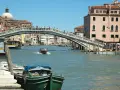 Un motoscafo naviga sotto uno dei ponti di Venezia, con edifici storici e gente che passeggia.