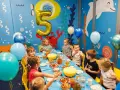 Dzieci świętują 5 urodziny przy stole pełnym przekąsek. Złoty balon w kształcie cyfry 5 unosi się nad stołem.