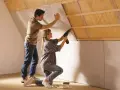 Un couple installe des plaques de plâtre sur un mur mansardé, préparant la surface avant un enduit à la chaux.