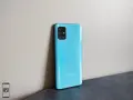 Błękitny Samsung A51 z aparatem w kwadratowej ramce, oparty o jasną ścianę.