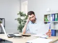 Zmartwiony mężczyzna z ręką na skroni analizuje dokumenty, być może nakaz zapłaty. Na biurku laptop, tablet i kalkulator.