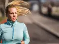 Kvinna med blont hår i hästsvans springer utomhus, lyssnar på musik med hörlurar. Hon är fokuserad på att springa 5 km tid.