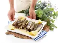 Un plato blanco con navajas a la plancha, limón y perejil. Unas manos sostienen el plato, decorado con eucalipto y flores moradas.