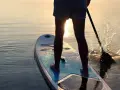 Person lernt SUP auf ruhigem Wasser bei Sonnenuntergang. Das Paddel spritzt Wasser auf.