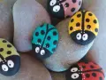 Seis piedras pintadas fáciles con mariquitas de colores: roja, amarilla, verde, rosa y turquesa, descansan sobre piedras grises.