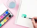Une main tient un pinceau pour créer une tache verte sur un carnet, à côté d'une palette d'aquarelle et d'un verre d'eau.