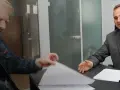 Dwóch mężczyzn przy biurku przegląda dokumenty, jeden w garniturze, drugi w dżinsowej kurtce.