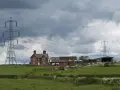 Wieś z domem i budynkami gospodarczymi, nad którymi wisi linia wysokiego napięcia.