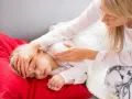 Matka troskliwie opiekuje się dzieckiem, które leży na czerwonej poduszce. Dziecko ma zamknięte oczy, a matka delikatnie dotyka jego czoła, sprawdzając, czy nie ma gorączki. To obrazek pokazujący troskę o dziecko podczas jelitówki.