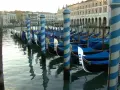 Una fila di imbarcazioni tipiche veneziane, le gondole, ormeggiate lungo un canale, con i loro caratteristici pali a strisce blu e bianche.