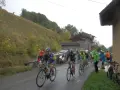 Ciclisti in gara su una strada bagnata, con una **forcella alta** che indica la salita. Il paesaggio è autunnale, con alberi e un pendio erboso.