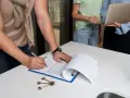 Hombre firma contrato, llaves en mesa. Se discute cuanto cuesta registrar una propiedad con personas al fondo.