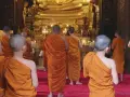 A group of Buddhist monks, another word for ascetics, walk towards a large golden Buddha statue in a temple.