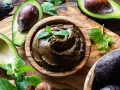 Crema di avocado vellutata in ciotola di legno, circondata da avocado freschi e foglie di menta.