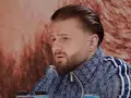 Amadeusz Ferrari w niebieskiej bluzie z białymi paskami, z charakterystyczną fryzurą i brodą, siedzi przed ekranem.
