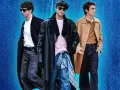 Tre uomini con un outfit elegante con jeans. Uno indossa un cappotto nero, camicia bianca e jeans grigi. Un altro un cappotto in pelle nero, maglione rosa e jeans blu. Il terzo un cappotto cammello, maglione marrone e jeans scuri.