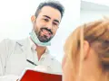 Uśmiechnięty dentysta w białym fartuchu i masce na szyi rozmawia z pacjentką, trzymając notatnik.