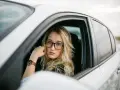 Blondynka w okularach siedzi za kierownicą białego samochodu. Pokazuje, jak nosić okulary, by wyglądać stylowo i pewnie.