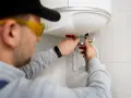 Instalator w okularach ochronnych podłącza bojler elektryczny, pracując przy rurach.