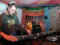 DJ Maceo Wyro w akcji, miksuje muzykę w klubie. W tle widać innych DJ-ów i kolorowe oświetlenie.