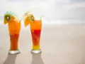 Dwa drinki Malibu z sokiem pomarańczowym, ozdobione plasterkami kiwi i pomarańczy, stoją na piaszczystej plaży.