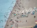 Tłum ludzi odpoczywa na kamienistej plaży w Finike. Niektórzy kąpią się w morzu, inni opalają się pod parasolami.