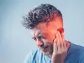 Mężczyzna z grymasem bólu na twarzy, trzymający się za ucho. Czerwony blask wokół ucha sugeruje **efekt zatkanego ucha** lub zapalenie.