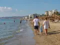 Rodzina spaceruje po piaszczystej plaży w Durrës. W tle widać morze, budynki i parasole plażowe.