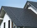 Ciemnoszara dachówka, czy to dachówka betonowa, czy ceramiczna, pokrywa dwa spadziste dachy domu pod błękitnym niebem.