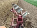 Czerwony pług z kilkoma lemieszami pracuje na polu, odwracając ziemię. Trwa regulacja pługa, przygotowując pole do siewu.