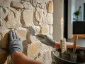 Mains gantées travaillant sur un mur en pierres naturelles, appliquant du mortier avec une truelle. Le mur qui s'effrite retrouve sa beauté.
