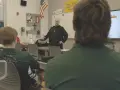 A monastery leader in black robes addresses students in a classroom.