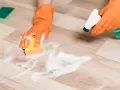 Hands in orange gloves spray cleaner and scrub a vinyl plank floor with a sponge, showing how to clean vinyl plank flooring.