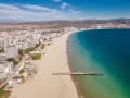 Słoneczny brzeg we wrześniu. Długie molo wchodzi w turkusowe morze, a za nim rozciąga się piaszczysta plaża i nadmorskie miasto.