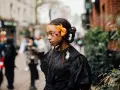 Ragazza con trecce davanti e capelli sciolti, ornata da un fiore arancione e makeup audace. Indossa un abito nero con dettagli voluminosi.