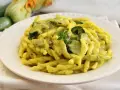 Piatto di pasta con zucchine e zafferano, guarnito con un rametto di erbe aromatiche. Sullo sfondo zucchine fresche e fiori di zucca.