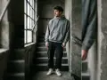 Giovane uomo con felpa oversize e pantaloni cargo scuri appoggiato a un muro di cemento, con scale e finestra sullo sfondo.