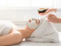 A spa technician applies a white mask to a woman's face. She's wrapped in a towel, enjoying a relaxing facial or massage first.