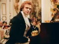 Piotr Adamczyk w historycznej scenie przy fortepianie, otoczony gośćmi w strojach z epoki. Jego role w filmach, serialach i programach zapadają w pamięć.