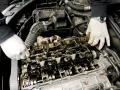 Mechanik w białych rękawiczkach sprawdza wałek rozrządu, szukając objawów zużycia. Widoczne są elementy silnika i przewody.