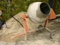 Betoniarka 200l czeka na piasek i cement. Ile cementu na betoniarkę 200l? Obok łopata i kapelusz.