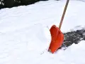 Pomarańczowa łopata do śniegu wbita w zaspę na ośnieżonym chodniku.