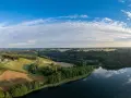 Malownicze **lasy w Polsce** otaczają spokojne jezioro. W oddali widać wieś z czerwonymi dachami, pola i pagórki.
