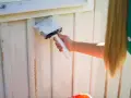 En person med långt blont hår målar en vit träfasad med en bred pensel. Det är dags att måla av huset för sommaren.