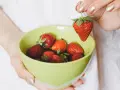 Hände halten eine hellgrüne Schüssel mit frischen Erdbeeren. Eine Erdbeere wird über der Schüssel gehalten, bereit zum Waschen.